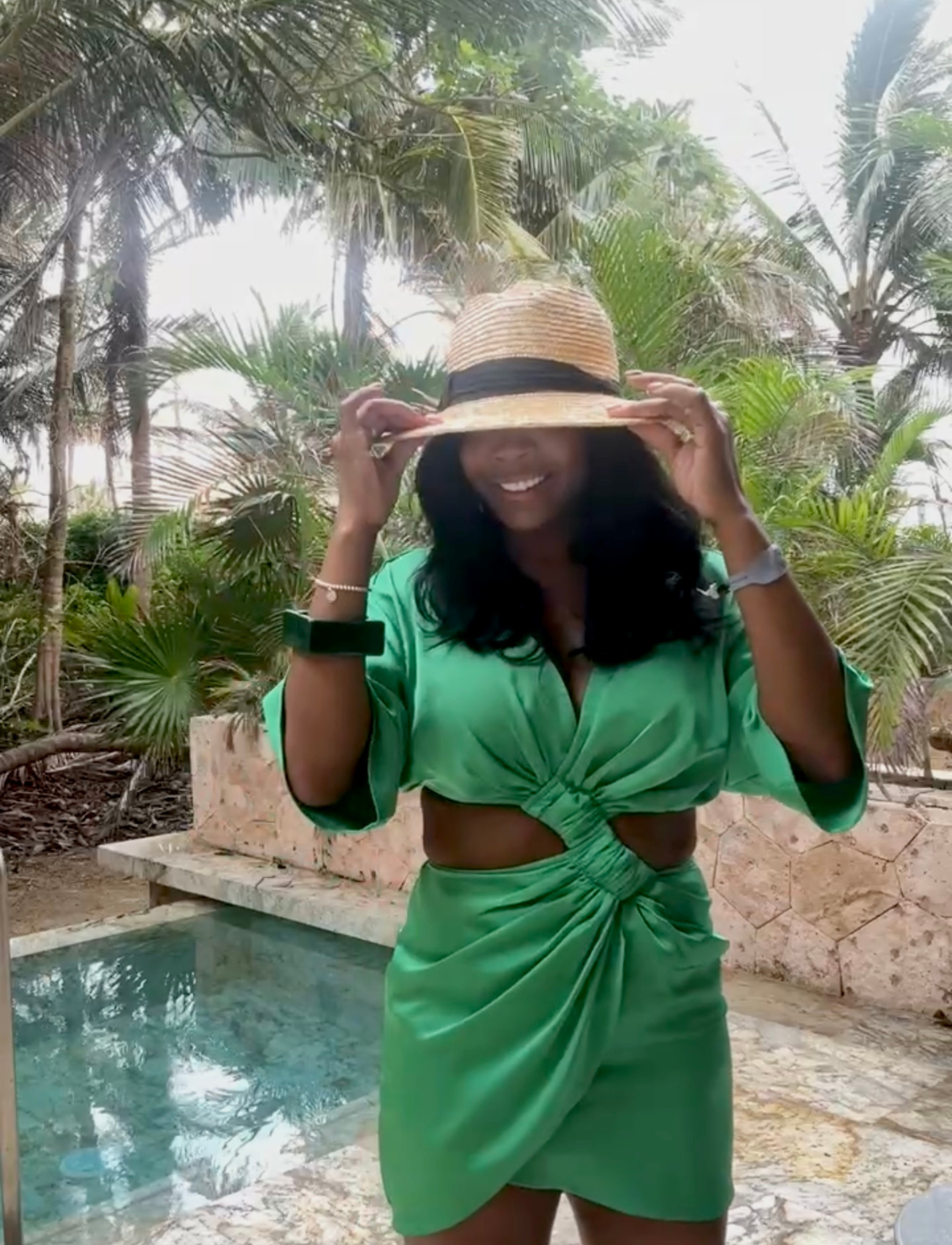 Woman in a green outfit and hat by a pool with palm trees in the background