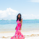 Woman in a pink floral dress standing on a beach with clear blue water and sky.